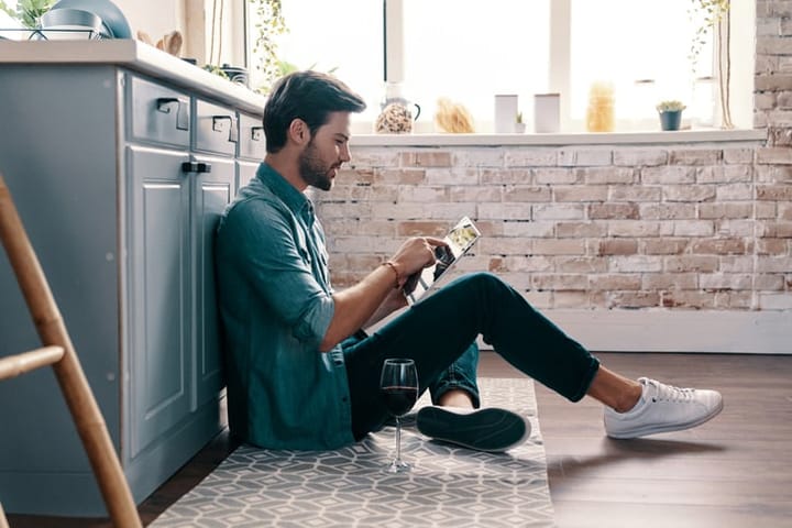 Handsome young man in casual wear using digital tablet and drinking wine while sitting on the kitchen floor