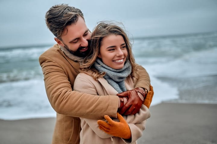 Beautiful married couple hugging on the seashore, wearing a coat in the cold season.