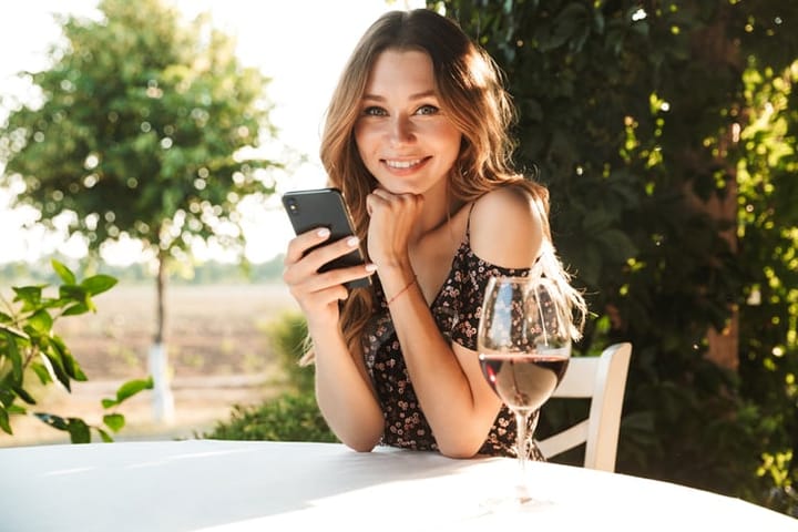 Image of young lady sitting in cafe outdors in park holding glass drinking wine chatting by mobile phone. Looking camera.