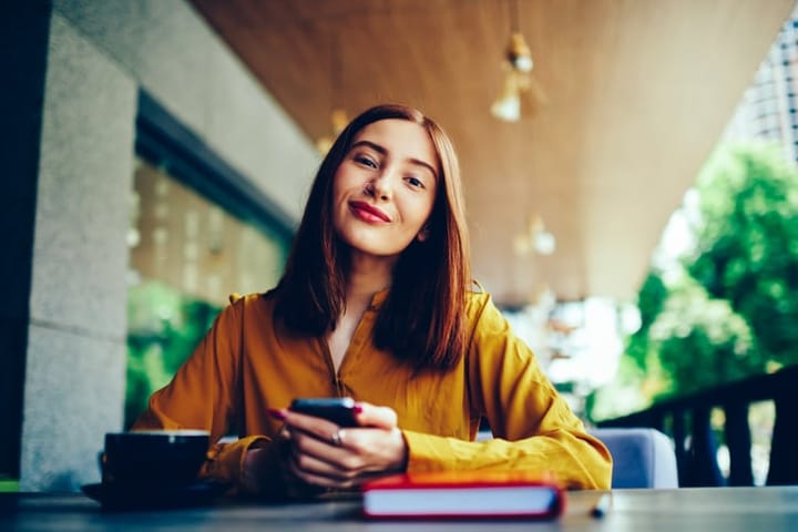 Half length portrait of positive charming female student looking at camera during spending leisure time with modern device and cup of coffee.Young pretty hipster girl sending messages to friends