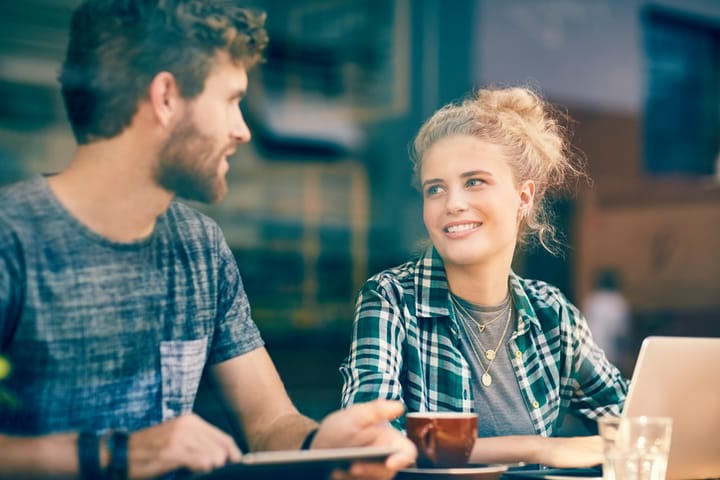 Smiling woman looking at man in cafe. Male and female customers are using laptop and digital tablet. They are seen through window of coffee house.