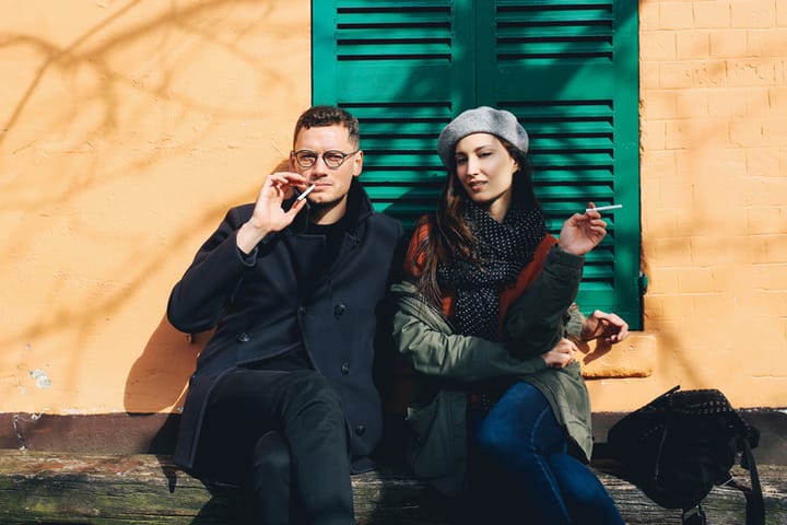 Vintage toned image of a young modern couple sitting and having a cigarette on the streets of Paris, near Montmartre, on a bright, spring day. He is wearing a black coat and glasses and the girl is in an olive green parka jacket and a scarf, wearing a typical French beret hat. Taken with a large aperture prime lens with added film emulation color grading.