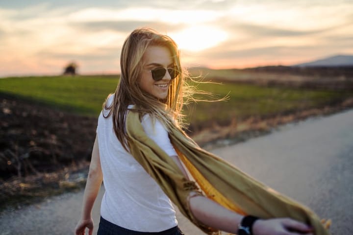 Young woman wearing sunglasses and a yellow scarf walking on the road on a sunny windy spring day