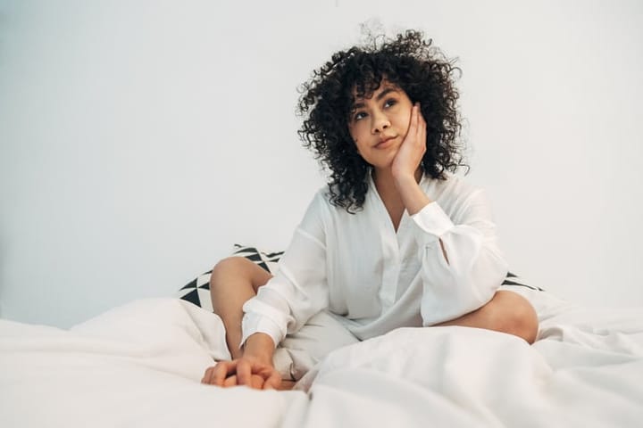 Pensive young woman sitting on bed resting face on hand.