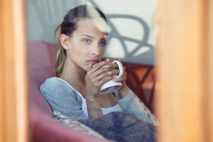 Shot of serious young woman looking through the window while drinking coffee on the sofa at home.