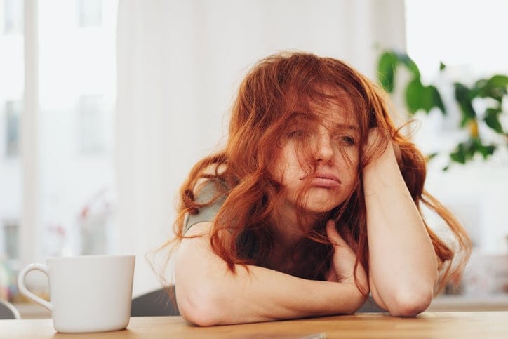 Red-haired girl sitting at the table indoors with boring face and looking away through messy hair, leaning on her elbow
