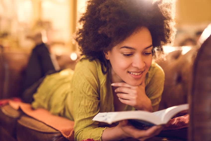 Smiling African American woman relaxing on sofa at home and reading a book.