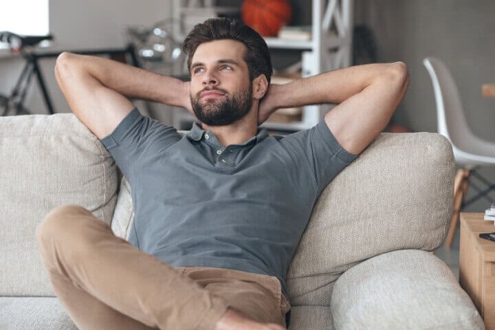 Handsome young man keeping hands behind his head and looking thoughtful while sitting on the couch at home