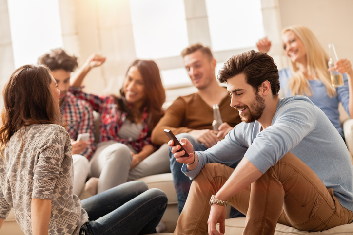Picture of a young smiling man using his smartphone with his friends in the background. The men and the women are drinking and cheering and all having a great time. They are sitting on beige sofas in a room with warm light and big windows.