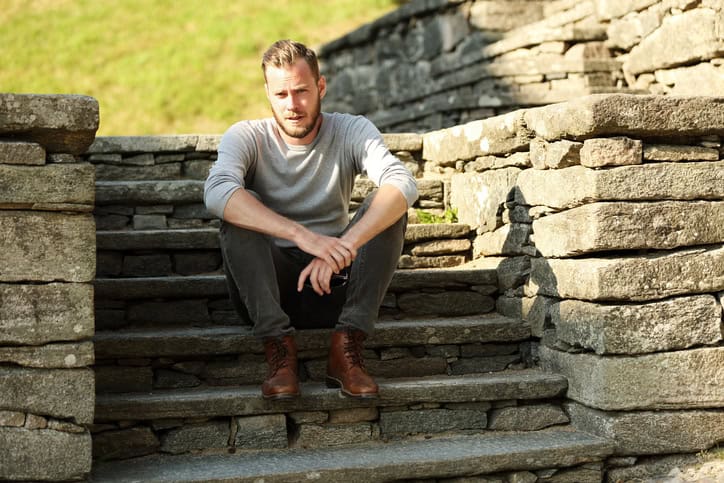 Man in his 20s wearing a grey shirt and jeans, sitting down outside on a set of steps on a sunny summer day.