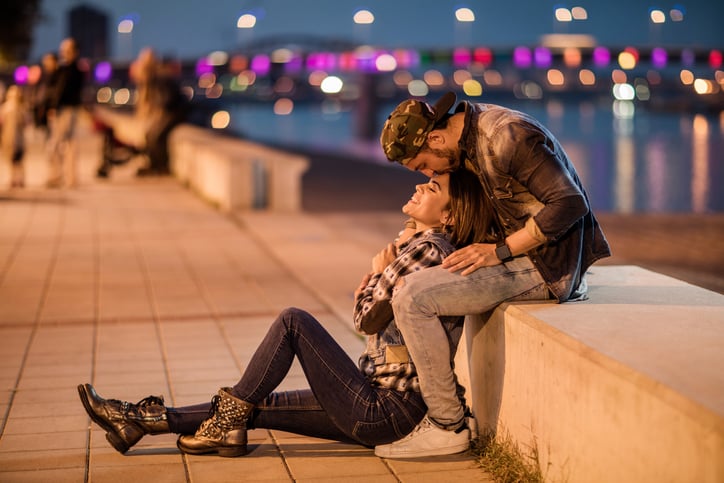 Loving couple relaxing on a riverside during the night while man is kissing his girlfriend on a forehead.