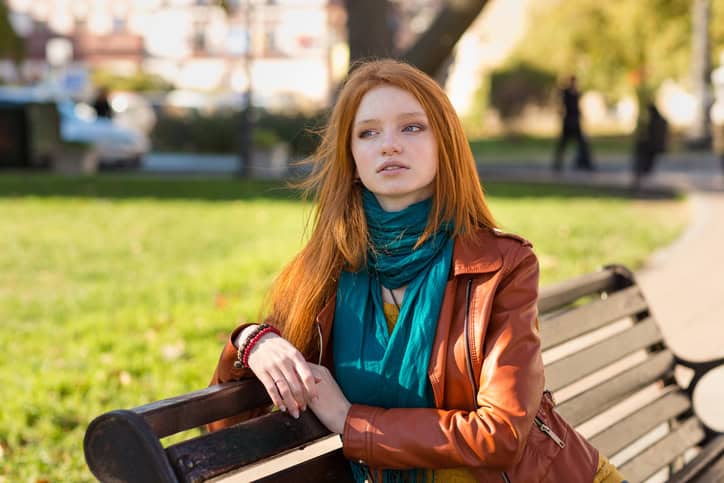 Young attractive pensive woman with beautiful red hair in leather jacket sitting on bench in the park