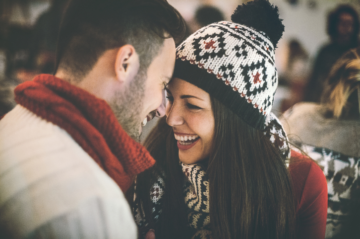 Closeup of happy mid 20's couple having fun at cosy coffee place on winter night. They are sitting face to face by a large window. Both wearing warm casual clothing. Blurry Christmas lights and decoration in background. Tilt shot,toned image.