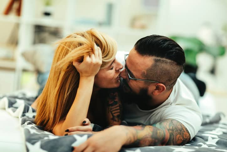 Young Tattooed Couple laying on front on sofa in their living room. Selective focus on their faces. They are enjoying spending time together.Kkissing with eyes closed.