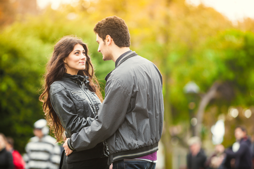 Young turkish couple is interacting while meeting in the city. The girl is looking up to her boyfriend's eyes with expectations and hope. She is shy and cute. He has his arms around her, admiring her. Beautiful autumn colour trees and unrecognisable people walking in the background. The couple stands out from the crowd.