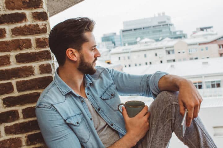 Young man at home balcony using smart phone and drinking coffee. He is happy and cheerful. Enjoying in sunny morning.