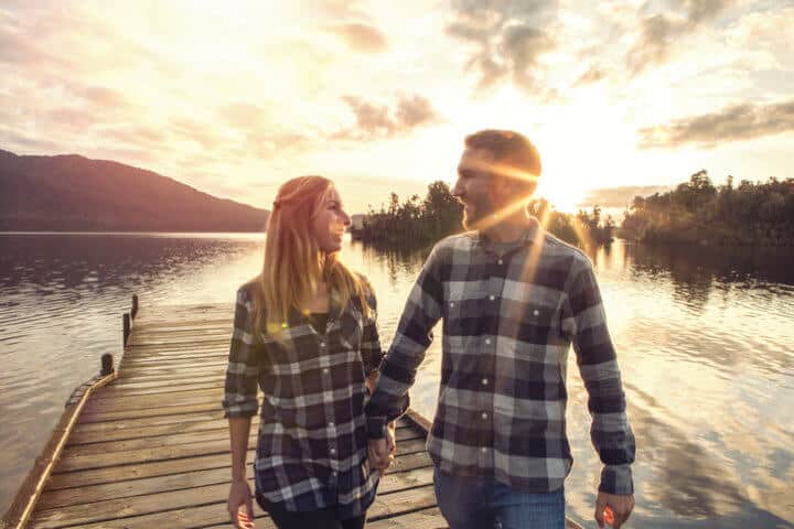 Cheerful young couple walking hand to hand on wooden pier above lake at dusk. South Island, New Zealand.
