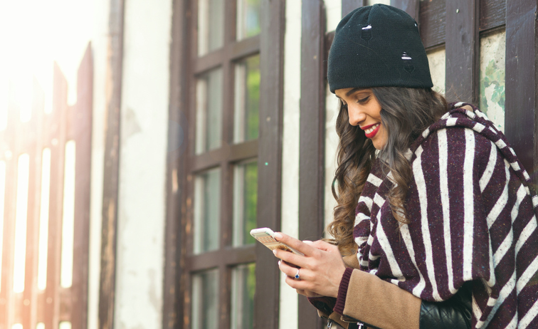 Picture of a young smiling woman sitting under the three on a cold winter day and using her smatphone
