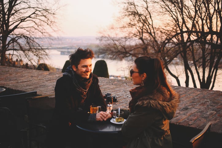 Young couple sitting at the outdoors coffee shop in the park. A couple in love holding hands, drinking tea, and smiling at each other. Young man and young woman facing each other amongst the park trees on a sunny winter afternoon.