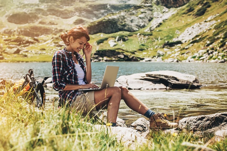 Young woman sitting at a lake side in the mountain and working on a laptop.