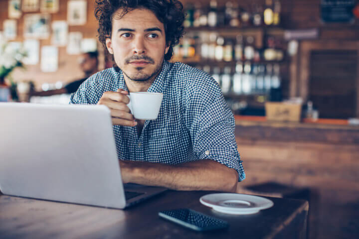 Young man sitting in bistro with a laptop in front of him, looking away.