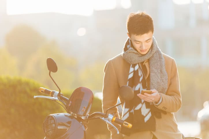 young asian man with mobile phone on motorcycle on road in modern city