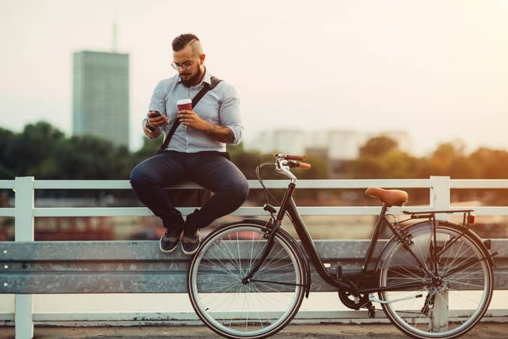 Young Businessman Sitting on fence in the city, using his smart phone and drinking coffee to go. He has tattoos and hipster haircut. Riding city styles bicycle to and from work.