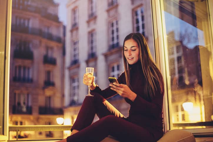 A young woman texting on the phone and drinking wine at home.