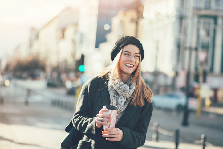 Smiling young woman in warm clothing walking under the sunlight at city scene, with copy space