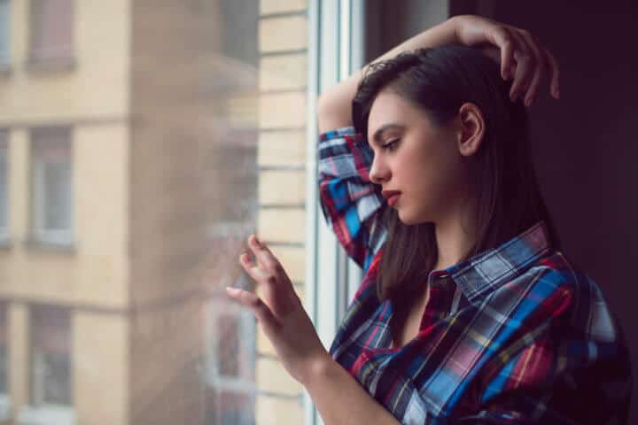 Beautiful young woman standing and leaning on the window pensive and touching the glass