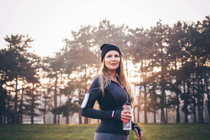 Young woman jogging outside in sunny autumn forest. She took a break to rest and drink water. Looking at camera and smiling. The sun is shining in the background .