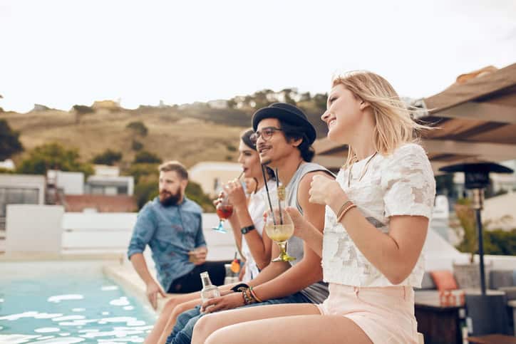 Happy young friends sitting by the pool having cocktail. Young people relaxing by the swimming pool with their feet in water during a party. Men and women enjoying rooftop party.