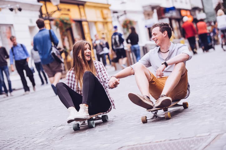 Portrait of happy young couple sitting on longboards holding hands. Man and woman skating and having fun on city street.