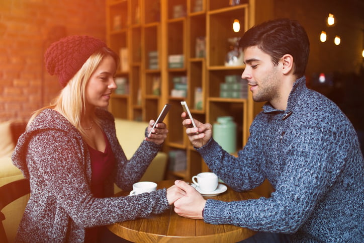 Young couple is sitting in a coffee shop holding hands and text messaging on the mobile phones at the same time without