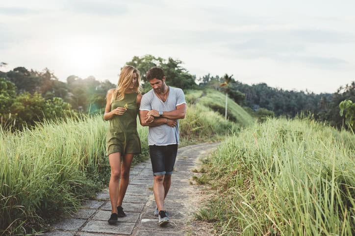 Young couple holding hands and walking through pathway in rural field. Man and woman walking along tall grass field.