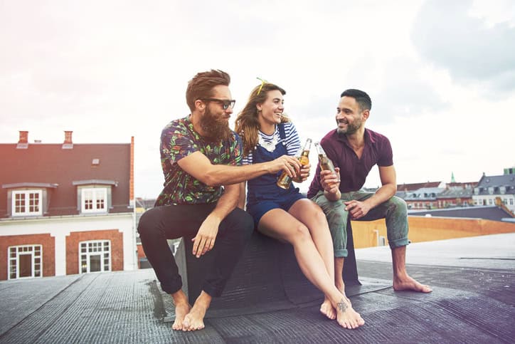 Cheerful group of barefoot adults drinking beer on roof outdoors with copy space in sky