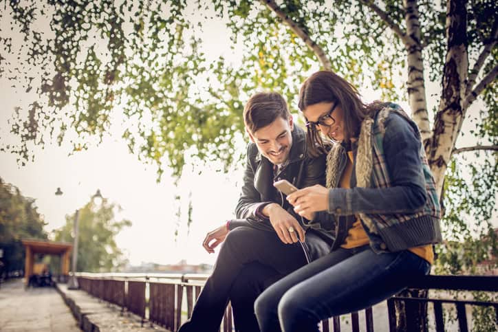Young couple sitting on a fence in the park, looking at the mobile phone, enjoying the sunny spring day outdoors.