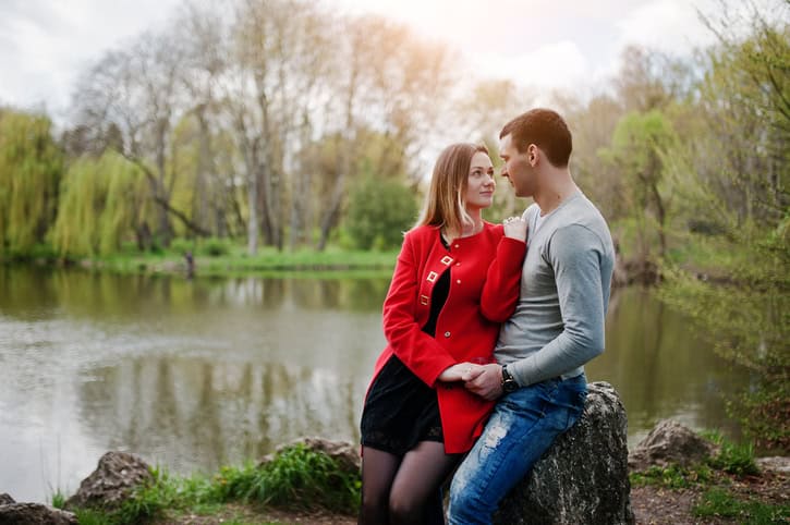 Happy loving couple sitting and hugging on stone at park background lake