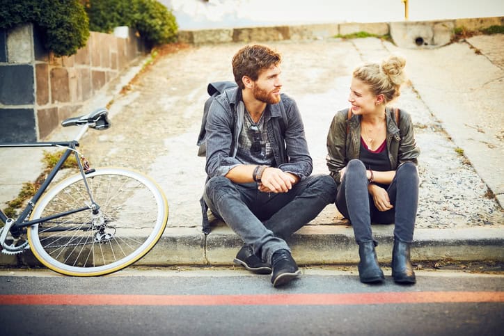 Young couple talking on sidewalk. Smiling male and female commuters are sitting at roadside. Full length of man and woman are casuals in city.