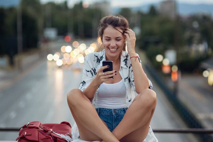 Girl looking at her mobile phone and laughing while sitting on a bridge