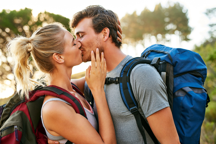 Loving male and female hikers are kissing in forest. Young affectionate couple is carrying backpack. They are wearing casuals in woodland.