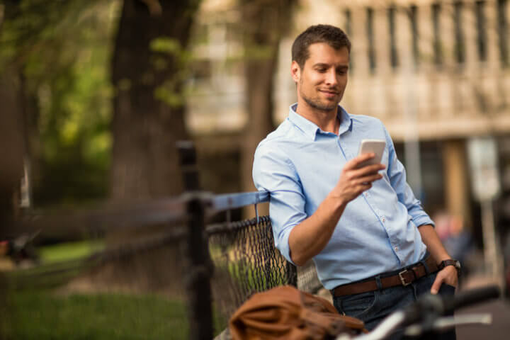Picture of a young business man who stopped on his bike to look at something peculiar on his smartphone. He is wearing a blue shirt with the sleeves rolled and he is leaning with his arm on a black fence. In the background are a green park and city buildings.