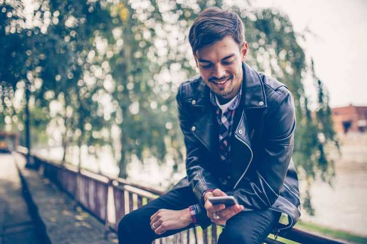 Young man using phone while sitting on a fence in city park