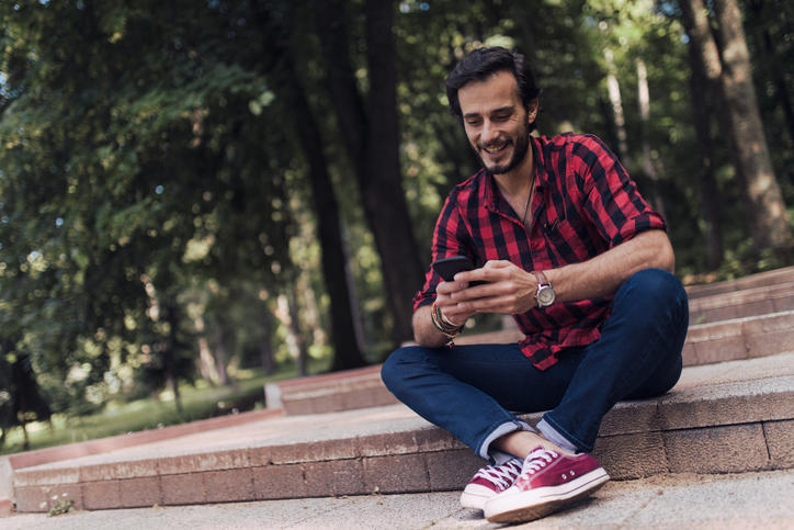 One man, sitting in park on steps, on a lovely day, using smart phone.