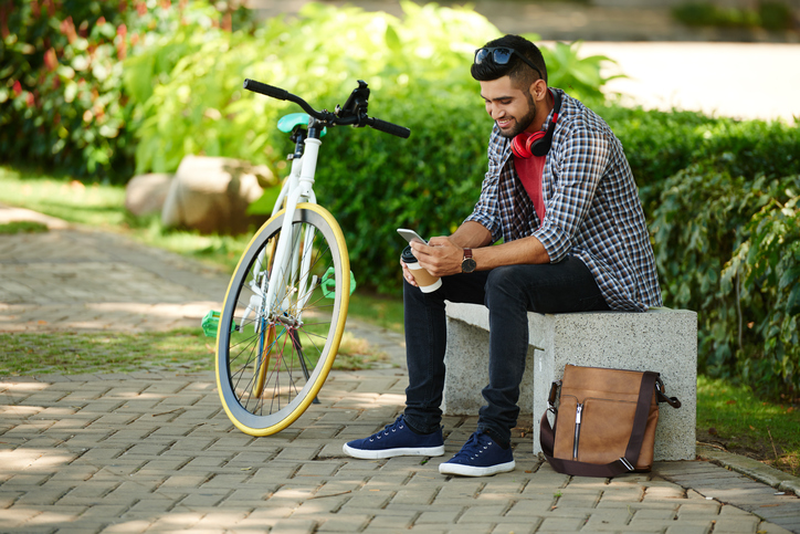 Smiling young man sitting on bench next to his bicycle and checking phone.