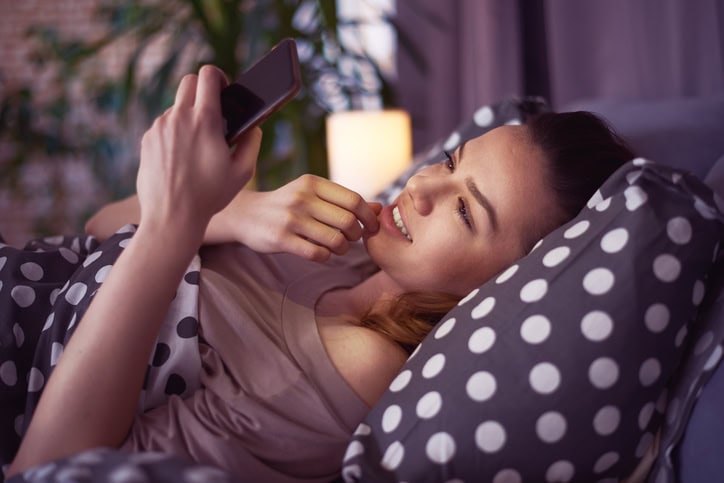 Girl laying down on bed in bedroom and using smart phone in the morning