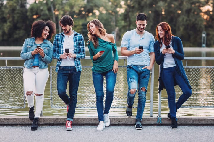 Multi-ethnic group of people standing in line outdoors and using mobile phones.