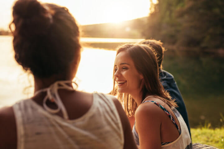 Portrait of happy young woman sitting by a lake with her friends. Young people enjoying a day at the lake.