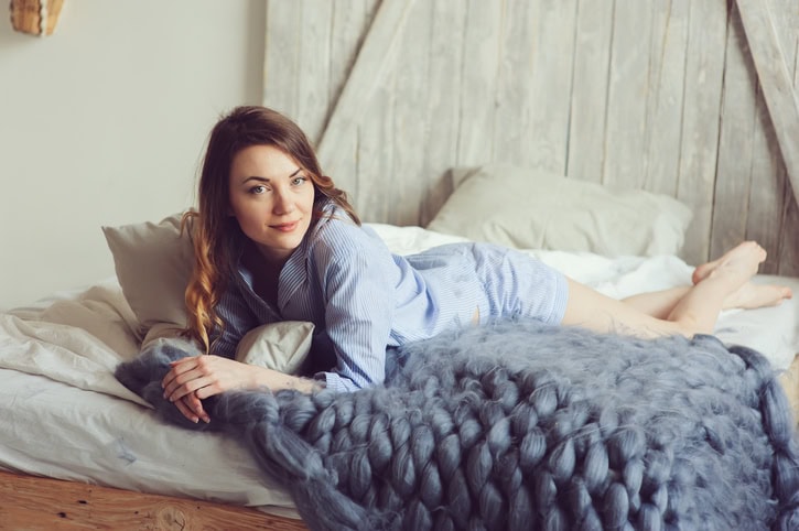 young woman in pajama wake up in the morning in cozy scandinavian bedroom and lying on bed with oversize knitted blanket. Casual lifestyle in modern interior