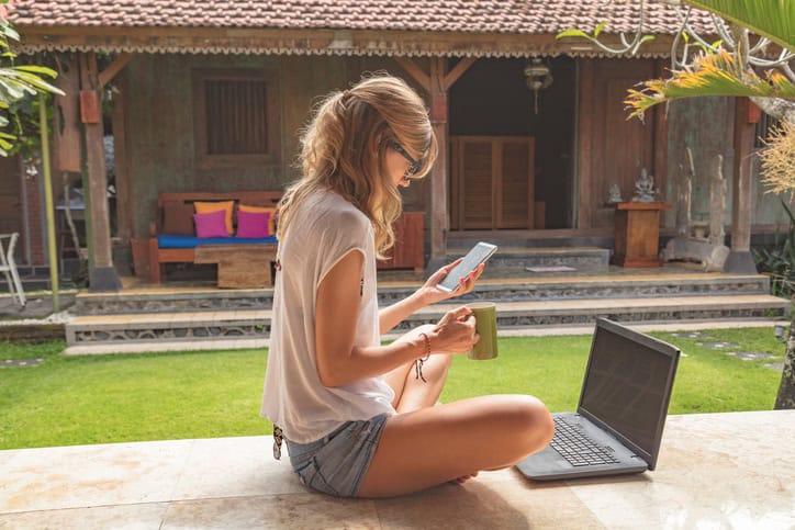 Girl holding cellphone, coffe mug with laptop on a garden porch.